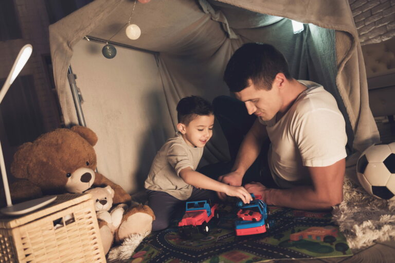 Father and son playing with cars in a blanket fort at night